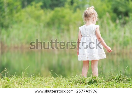 Two years old girl in light summer dress near green forest pond