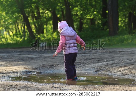 Curious little toddler girl is walking in summer puddle with gum red boots back view
