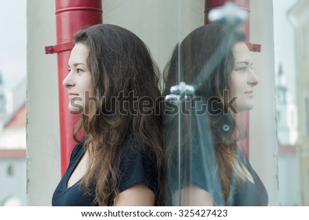 Glass portrait of looking forward feminine brunette girl is posing near display window with symmetry glass reflection