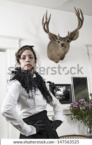 A woman in Edwardian style dress posing in the room below a stag\'s head hanging on the wall