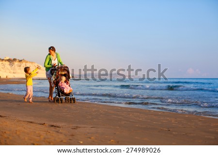 Single mother walking with her daughter and son, talking and pushing a stroller on a sandy beach in late summer, enjoying the evening chill. Family vacation, traveling with children concept.