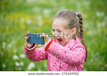 Little smiling and curious girl photographing with her smart phone, exploring nature and standing in a dandelion meadow. Active lifestyle, curiosity, pursuing a hobby, technology and kids concept.