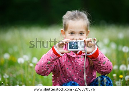 Little smiling girl proudly showing her photo on display, taken in a dandelion meadow. Active lifestyle, curiosity, pursuing a hobby, technology and kids concept.