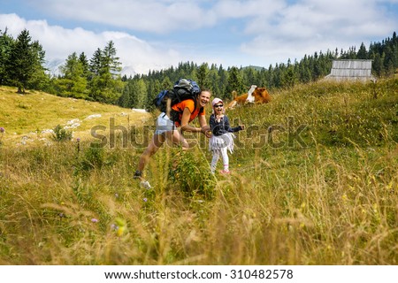 Mother tickling her daughter on mountain hike in the Alps, having fun, spending quality time together, with huts and cows in background. Active parenting, fun childhood and family bonding concept.