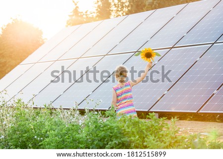 Young 6 year old blonde girl child standing in front of small solar panel farm in countryside. Renewable energy concept. Sun lens flare.