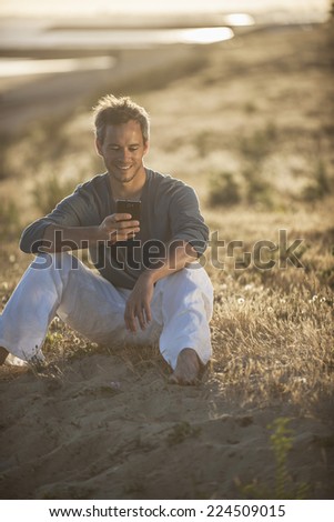 handsome man sitting on the beach at sunset and writing a sms on his phone