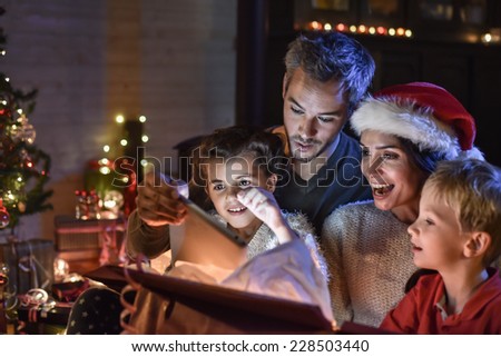 happy family finds a digital tablet in a gift on the Christmas night,  near the wood stove and lit Christmas tree in their living room, the mother wears a santa hat