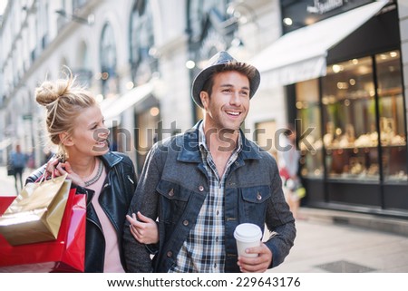 a trendy young couple walks in the city in autumn, the young woman wears a leather jacket , shopping bags at her arm and the man a cup of coffee in hand