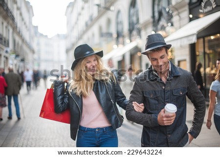 a trendy young couple  wearing hats walking in the city in autumn, the young woman wears a leather jacket , shopping bags at her arm and the man a cup of coffee in hand