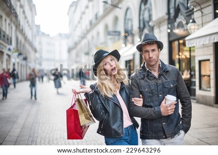 a trendy young couple  wearing hats walking in the city in autumn, the young woman wears a leather jacket , shopping bags at her arm and the man a cup of coffee in hand