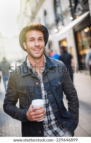 a smiling trendy guy with a hat, walking in the city a cup of coffee in hand