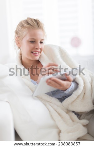 Beautiful blond woman using a digital tablet, lying on a couch wrapped in a white blanket