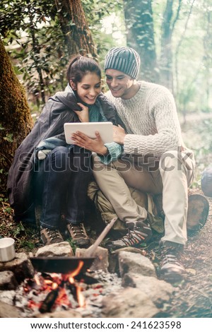 in the woods a couple sitting near a campfire using a digital tablet, she has a sleeping bag on the shoulders for warmth