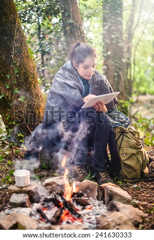 in the woods a young woman sitting near a campfire using a digital tablet, she has a sleeping bag on the shoulders for warmth
