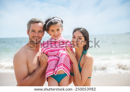 A young couple at the beach is posing in swimsuits with their daughter, Mom and dad are drying their six year old girl with a towel, family looking at the camera.
