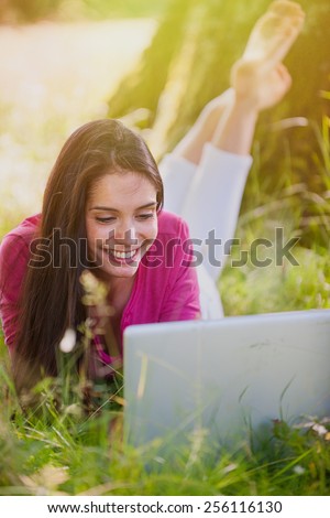A smiling woman in her 30\'s is lay down on the grass looking her computer. She is relaxing, enjoying the shadow of the tree in a sunny day.