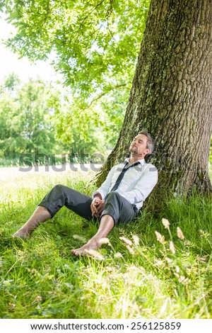 A nice looking man is sitting against a tree in the grass, looking like he is dreaming.  He is relaxing, enjoying the shadow of the tree in a sunny day.