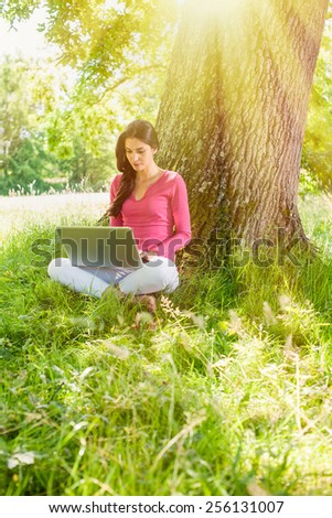 A nice looking woman is sitting against a tree in the grass, looking at her computer. She is relaxing, enjoying the shadow of the tree in a sunny day.