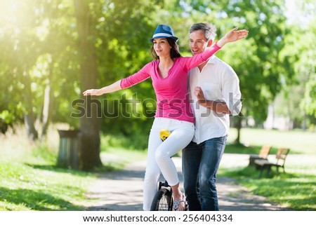 A lovely couple is having fun on a unicycle in the park. A beautiful woman is trying to ride the unicycle, while her grey hair boyfriend is holding her. They are wearing casual clothes.