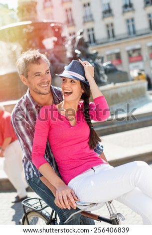 A smiling couple is riding a retro bike in the city center. The grey hair man is riding the bike while the woman is sitting on the handlebar. She is wearing a pink top and a blue hat.