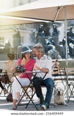 Portrait of a smiling couple eating ice cream on a terrace in the city. The grey hair man with a beard is in a white shirt. The woman is wearing a blue hat . A white dog is lay down behind them.