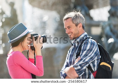 A loving couple is taking photos in the city center. . The woman is taking a closed picture of the grey hair man with a beard and a black backpack while he is smiling.