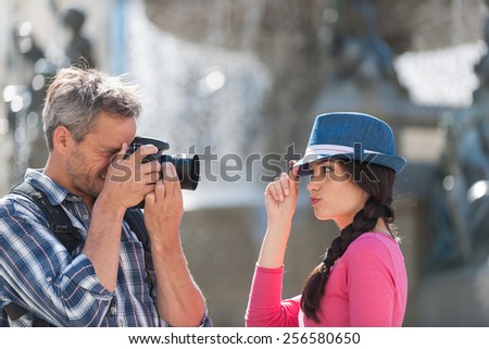 A loving couple is taking photos in the city center. . The grey hair man with a beard and a black backpack is taking a closed picture of the smiling woman with a blue hat and a pink top.