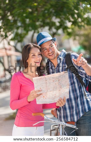 A young couple is looking for direction. A woman is holding a map, while a grey hair man is pointing some direction, holding a bike. The man with a beard is wearing a blue hat and a black backpack.