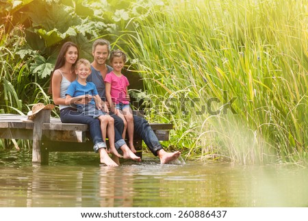 in summertime, portrait of an happy family sitting at the edge of a wood pontoon, feet in the river,  looking at the camera