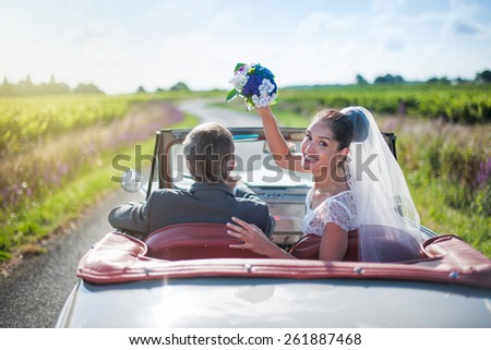 A newlywed couple is driving a retro car, the bride looks at the camera and throws her bouquet, rear view