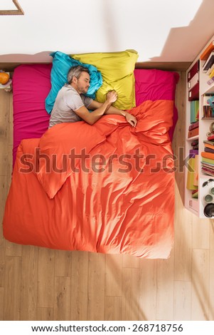 top view of a man lying in bed at bright colors, it has gray hair, a library is next to his bed in his sunny apartment