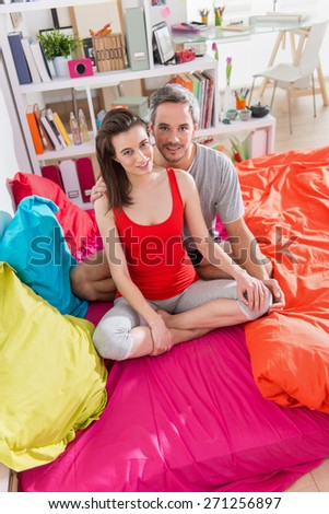 Looking at camera, a glamour couple in pajamas, sitting in bright colors\'s bed, man has gray hair, their apartment is modern and bright
