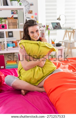 Looking at camera, a gorgeous young woman sitting in bed, clutching a green pillow in her arms,bedding is bright color, her sunny apartment is modern and bright