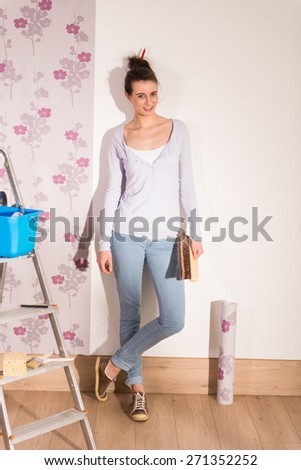 Looking at camera, a young woman posing wallpaper on the walls of her apartment, she is DIY dressed, her tools beside her