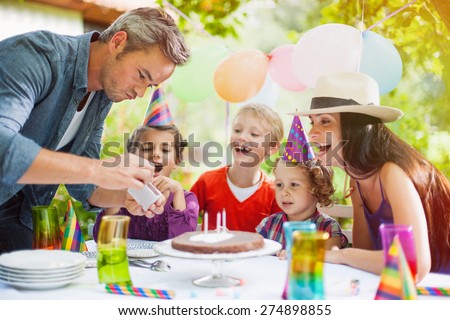 garden party with family for little girl\'s birthday, Dad tries to light the candles on the cake , the garden is decorated with balloons and colors are bright
