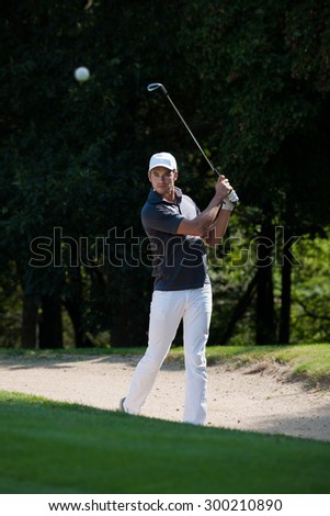 Handsome golfer in white pants and cap just hit the ball with his club on a nice green course. He just finished his swing and is holding his club up.