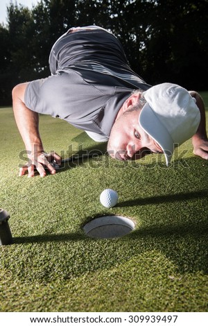 Grimacing golfer lying down on the green, blowing on his white ball to make it fall in the hole. Close up  on the ball and the man head with a beautiful forest in the background.