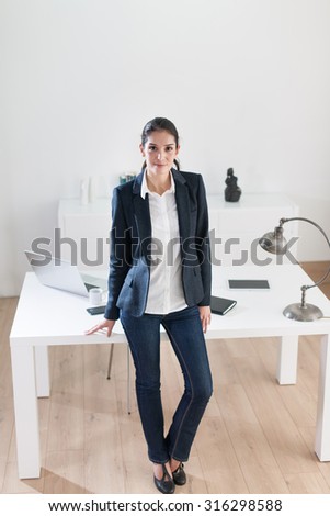 Top view of smiling successful creative businesswoman leaning against a white desk in a luminous office with wooden floor. She is looking at camera, wearing a black suit jacket and white shirt