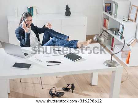Top view of a dark hair businesswoman sitting at her desk in a design luminous office. She is relaxing, talking on the phone, her barefoot on the desk, her high-heeled shoes are under the desk