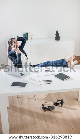 Top view of a dark hair businesswoman sitting at her desk in a luminous office. She is talking on the phone, touching her hair, her barefoot on the desk, her high-heeled shoes are under the desk