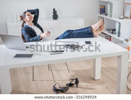 Portrait of a dark hair businesswoman sitting at her desk in a luminous office. She is scratching her head, her barefoot on the desk, holding her coffee mug. Her high-heeled shoes are under the desk