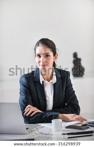 Close up of a smiling successful middle age businesswoman with dark hair sitting at her white desk in her office. She is wearing a black suit jacket, crossing her arms, her laptop next to her