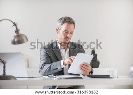 Close up of a smiling successful middle age businessman with grey hair and beard sitting at his white desk in his office He is working on his tablet with his laptop, glasses and smartphone next to him
