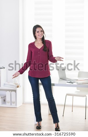 Portrait of a dark braided hair woman posturing in front of a white table, with a grey laptop, in a luminous open space. She is wearing casual clothes and looking at camera with her hands up