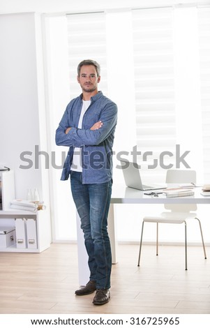 Portrait of a grey hair man with beard standing next a white table, with a grey laptop, in a luminous open space. He is wearing casual clothes and looking at camera with his arms crossed