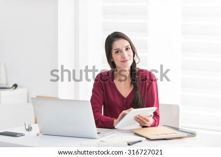 Portrait of a designer working home on new ideas A dark hair braided woman is sitting at a white table in casual clothes, she is looking at camera, holding her tablet and with her laptop next to her