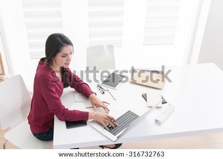 Top view of a designer working home on new ideas. A dark hair braided woman is sitting at a white table in casual clothes, she is working on her laptop and have some sketches on a notebook next to her