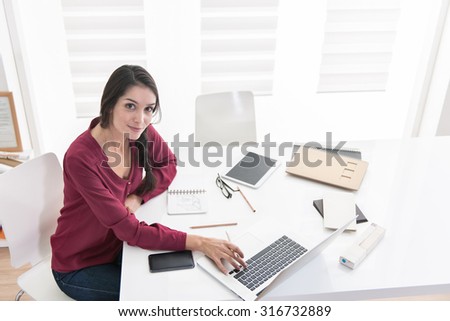 Top view of an architect working home on new ideas. A dark hair braided woman is sitting at a white table in casual clothes, she is looking at camera, her laptop and sketches on a notebook next to her