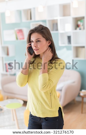 Focus on a woman in her 30's talking on her smartphone in her stylish vintage living room. The furniture and the wall are painted with pastel colors. She is wearing a yellow pull and black pants