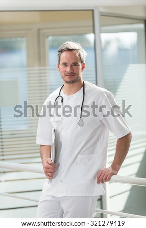 A nice grey hair doctor with beard is standing in his white coat, his stethoscope around his neck in front of hospital room with glass walls. He is looking at camera with holding patients charts
